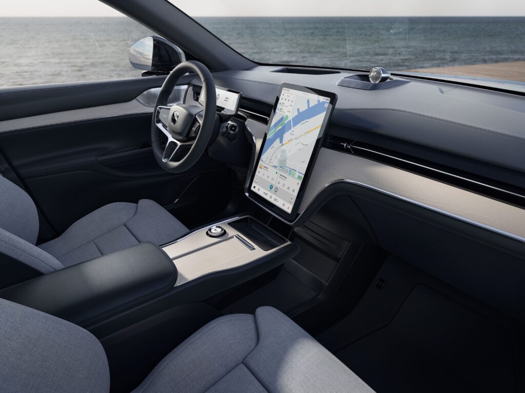 Volvo EX90:n sisätila kuvattuna sivulta, näkyvissä ohjauspyörä, keskikonsolin kosketusnäyttö ja etuistuimet, taustalla merimaisema.